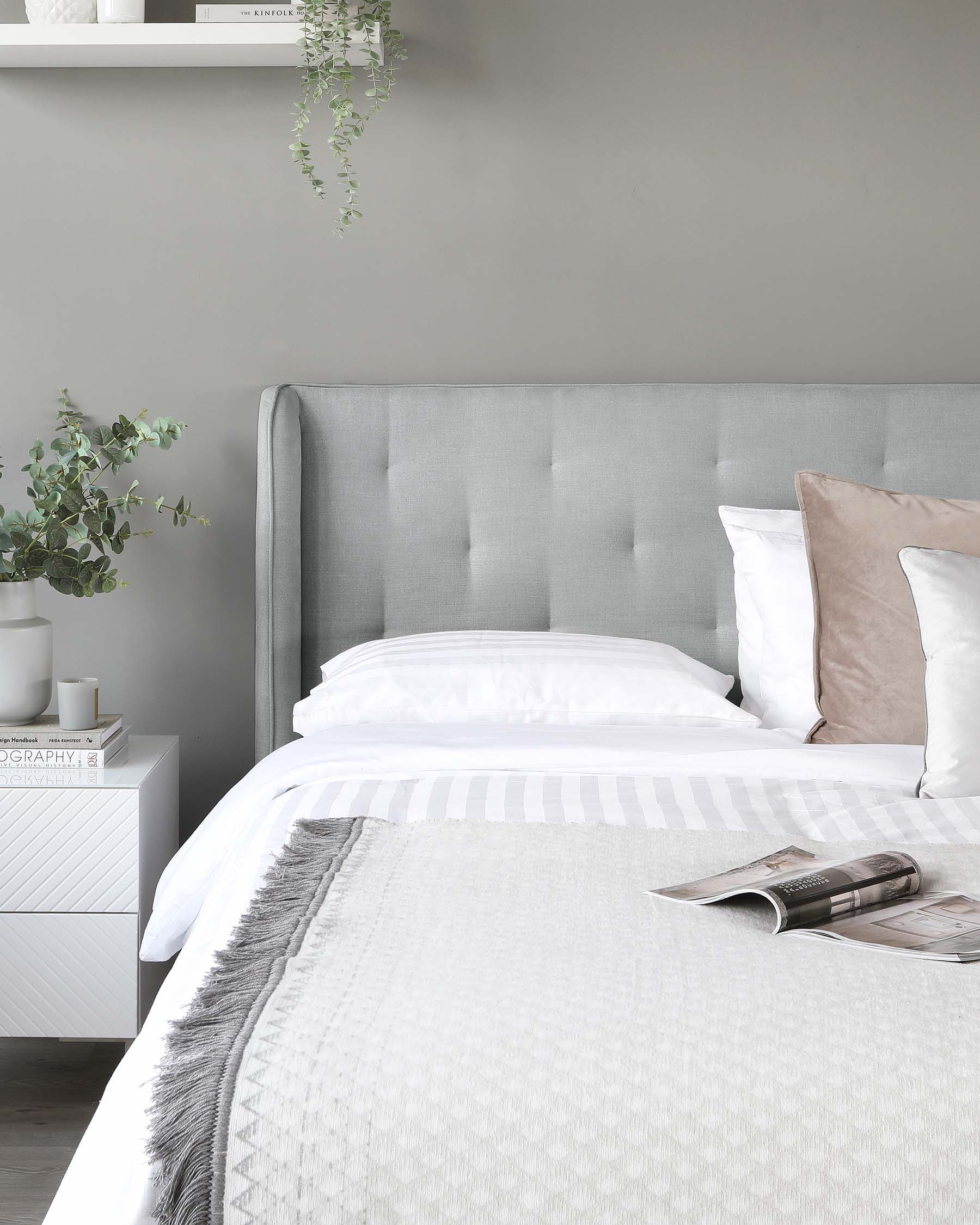 Elegant bedroom interior showcasing a contemporary upholstered grey tufted headboard, a pristine white textured bedding set complemented by soft beige and grey pillows. A white modern bedside table featuring clean lines and geometric-patterned facades, with a small potted plant and decorative items on top, enhances the serene and sophisticated ambiance of the space.