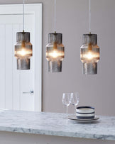 Three textured glass pendant lights hanging above a white marble countertop with two wine glasses and a stack of striped ceramic bowls.