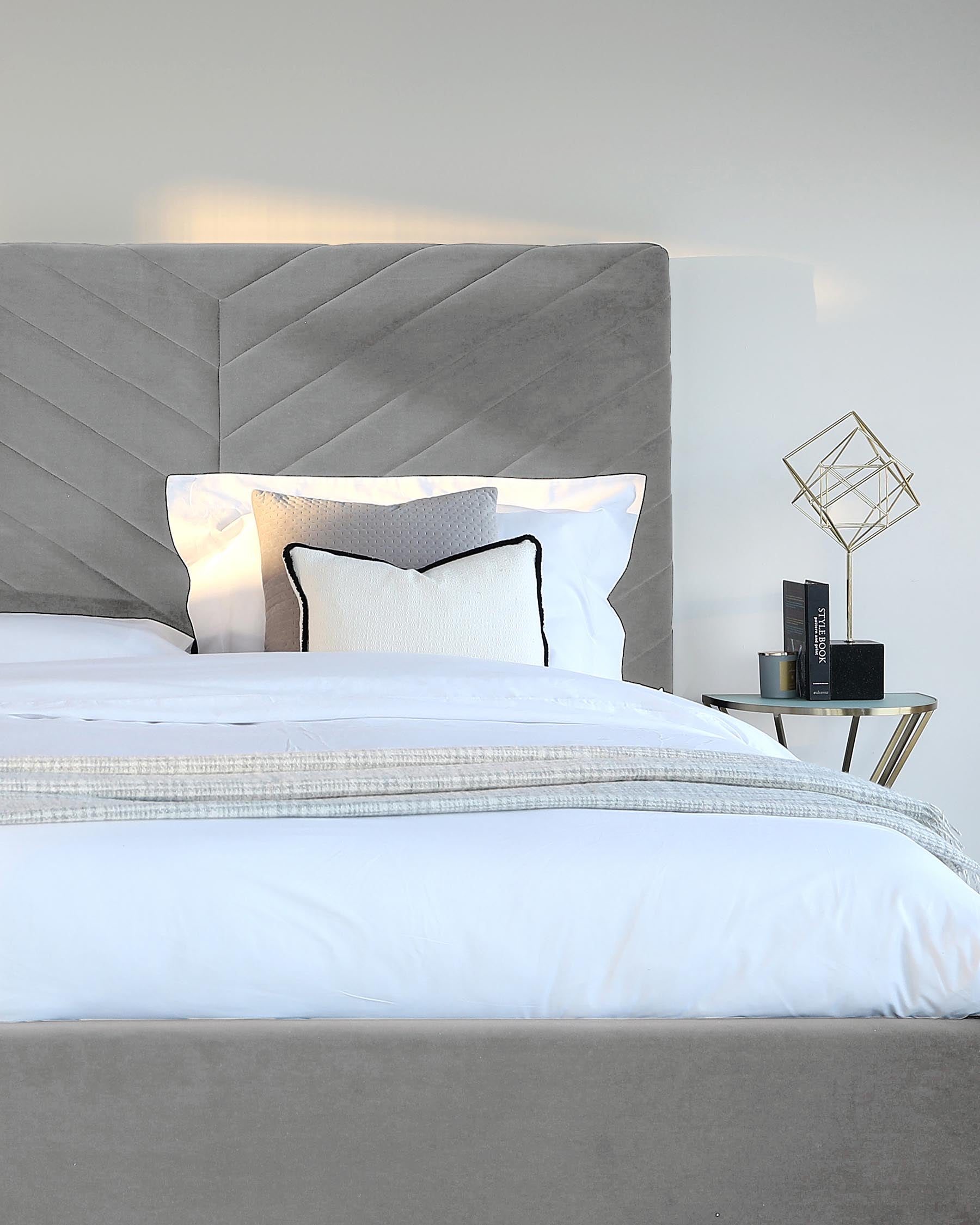 A contemporary grey upholstered bed with a large, plush headboard featuring a geometric chevron pattern. The bed is adorned with crisp white linens, a decorative grey throw, and several pillows, including one with a black piping trim. Beside the bed is a modern, minimalist gold-toned side table with a geometric metal table lamp and books.