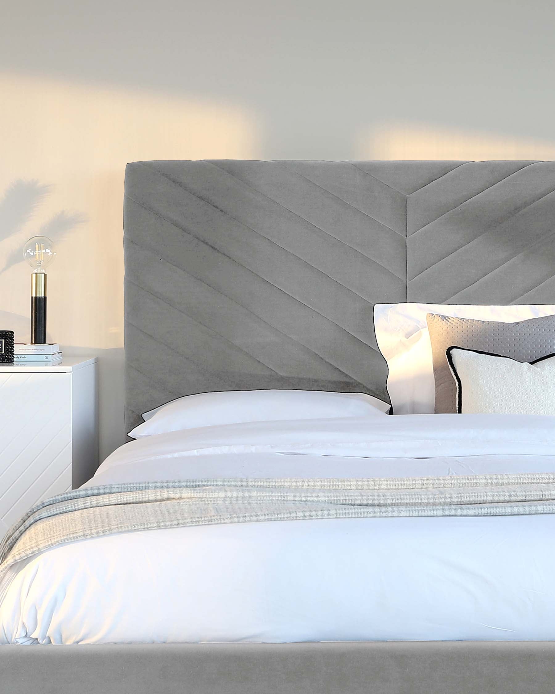 A modern bedroom featuring a large bed with an oversized grey upholstered headboard displaying a geometric chevron pattern. Crisp white bedding with an elegantly textured throw blankets the bed. A sleek white nightstand is partially visible to the side, topped with a small stack of books and a contemporary table lamp with a clear glass base and a white shade. The scene conveys a serene and luxurious atmosphere.