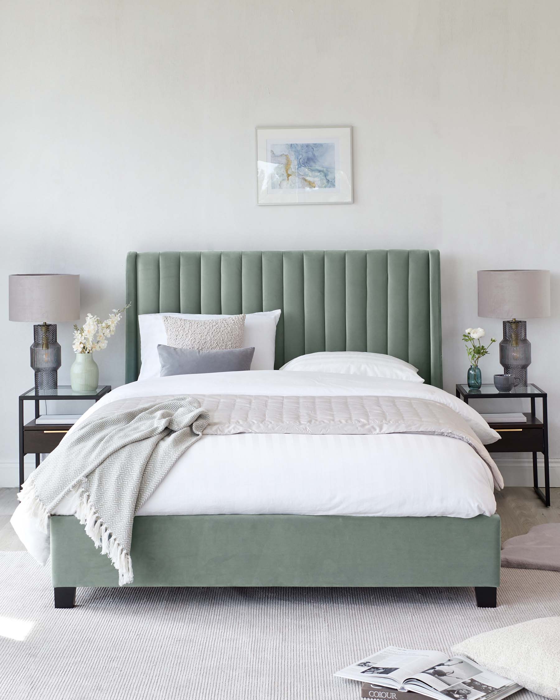 A contemporary bedroom featuring a plush, olive-green upholstered bed with a vertically channelled headboard. Flanking the bed are two black metal nightstands with glass tops, each holding a textured grey lamp and a vase with white flowers. A cosy throw blanket is casually draped at the foot of the bed on crisp white bedding.