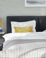 An elegant, contemporary bedroom featuring a plush, charcoal grey upholstered headboard which provides a sophisticated backdrop to a luxuriously dressed bed with crisp white linens and a textured grey throw. A mustard velvet accent pillow with decorative pompoms adds a pop of colour and a touch of playfulness to the setting.