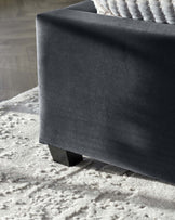 Close-up view of a dark upholstered furniture piece with a sleek modern design, featuring clean lines and a solid-colour fabric. The furniture has a plush appearance and is sitting on a shaggy, light-coloured rug that contrasts with the dark textile of the furniture. The visible leg is angular and black, suggesting a contemporary style.