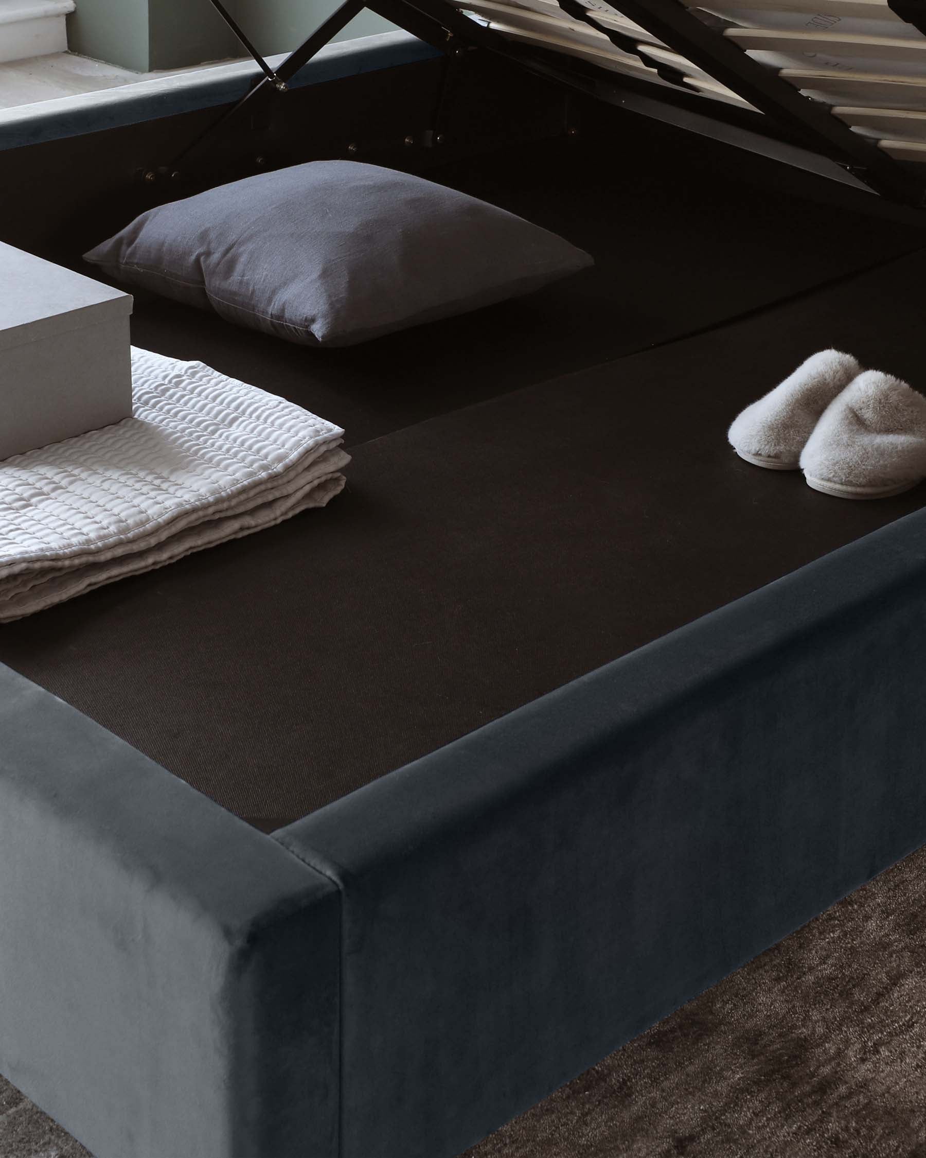 Modern minimalist-style upholstered storage bed in a dark blue velvet fabric with a lift-up mattress revealing storage space beneath, accompanied by a pair of grey fabric pillows and white textured bedding folded at the foot of the bed. Soft white slippers are placed on the dark fabric bed cover, evoking a cosy, inviting atmosphere.