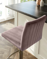 Elegant modern bar stool with a plush, mauve velvet seat featuring vertical channel stitching on the backrest, and a sleek metallic base with a footrest.