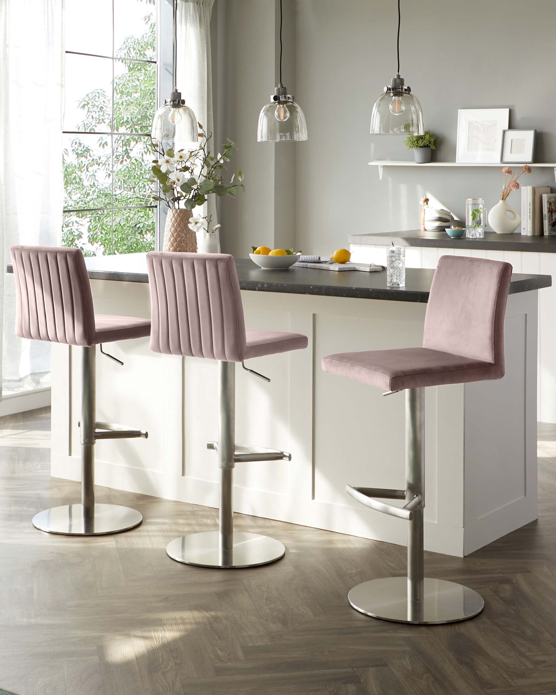 Three modern bar stools with pink velvet upholstery and vertical channel tufting on the backrest. Each stool features a chrome pedestal base with a footrest and adjustable height mechanism.