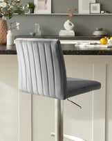 A modern bar stool with a grey upholstered seat featuring vertical channel stitching on the backrest. The stool has an adjustable-height chrome base with a footrest.