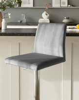 Modern bar stool with a sleek, grey velvet upholstered seat and backrest, paired with a minimalist metallic silver base, positioned against a kitchen counter with contemporary home decor in the background.