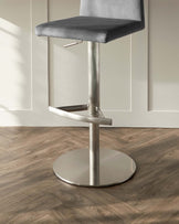 Modern bar stool with grey upholstery and a chrome pedestal base with footrest and flat circular bottom.