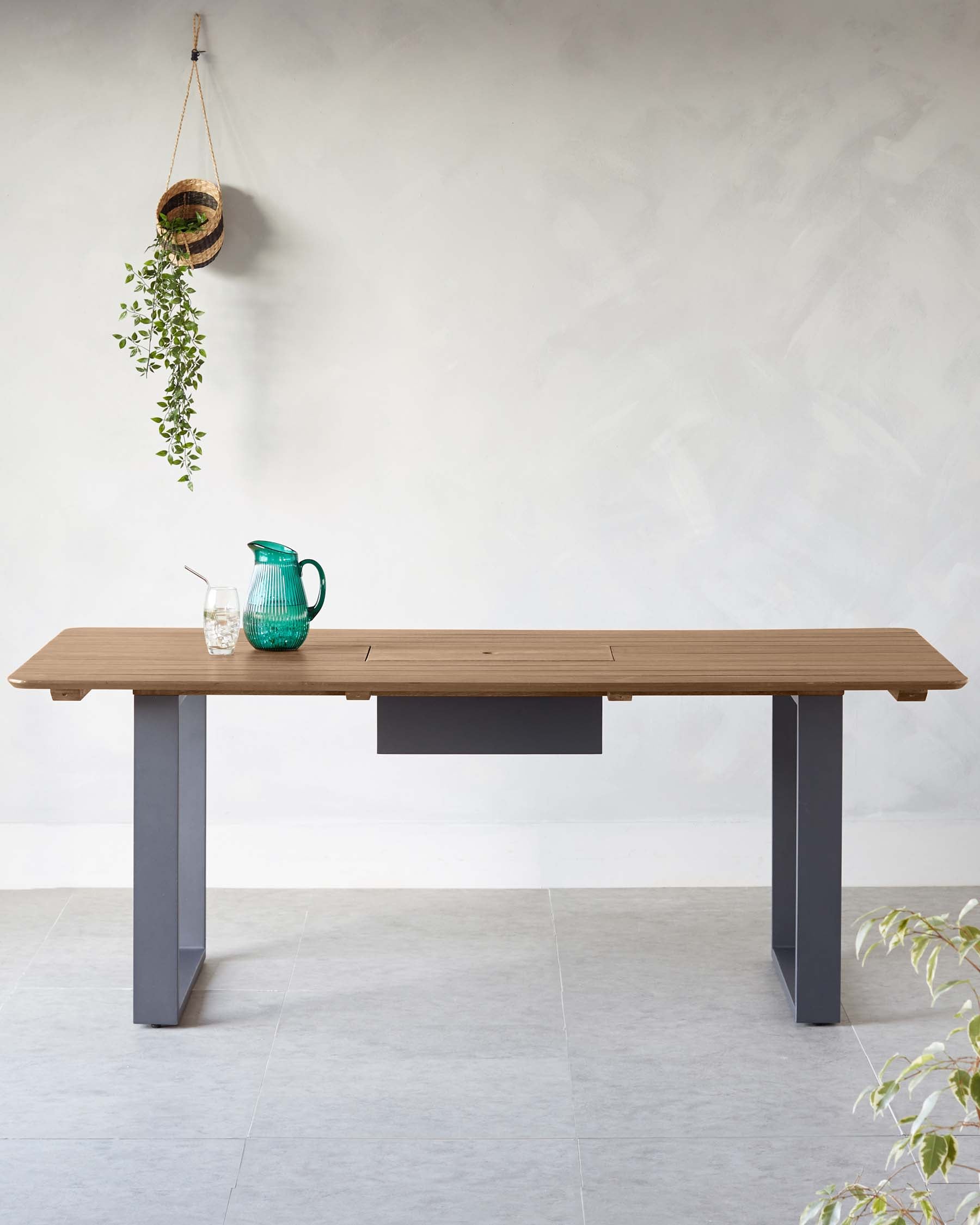 Modern wooden dining table with a light finish, sleek grey legs, and a minimalist drawer, paired with a green pitcher and glass.