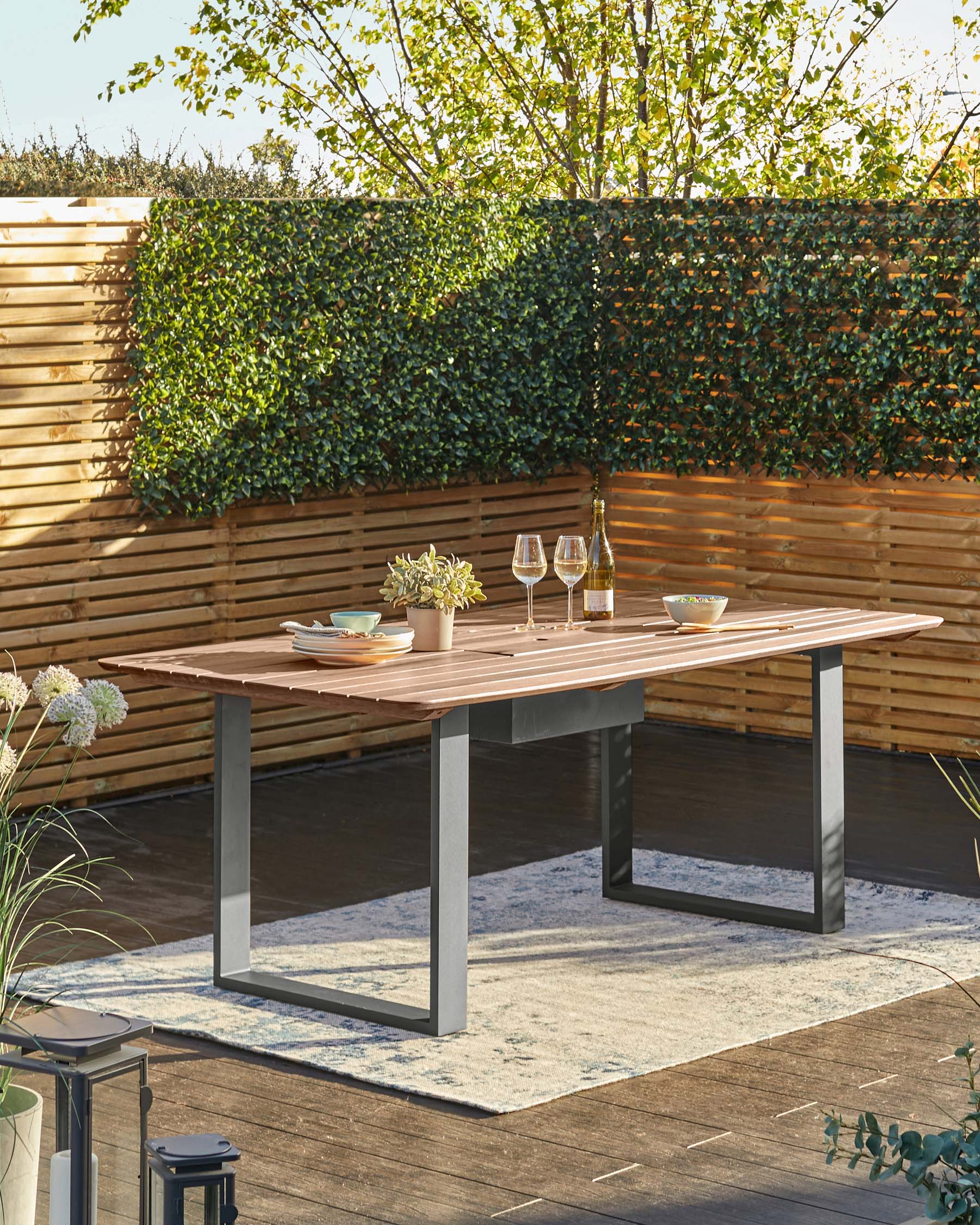 A modern outdoor dining table with a wooden top and metal legs, set with plates, glasses, and a decorative plant.