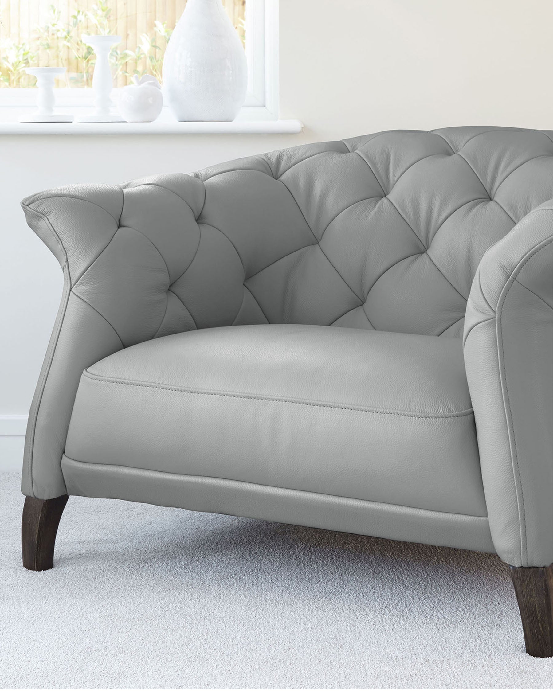 Elegant grey tufted loveseat with a curved backrest, plush seat cushion, and classic dark wooden legs set against a bright room with a white window sill displaying decorative vases.