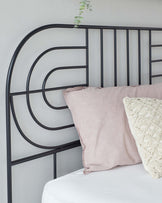 Modern minimalist black metal bed frame with a geometric headboard design, featuring straight vertical and curved horizontal bars. The bed is dressed with a plain white fitted sheet, a pink striped pillow, and an off-white textured throw pillow. There is a potted trailing plant atop the headboard adding a touch of greenery to the scene.