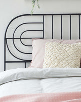 A modern metal bed frame with a geometric headboard pattern featuring circular and linear elements, in a black or dark finish. The bed is dressed with a textured white duvet cover, a pale pink sheet with a subtle pattern, and a decorative cream pillow with a knitted design. A hanging green plant adds a touch of nature to the scene.