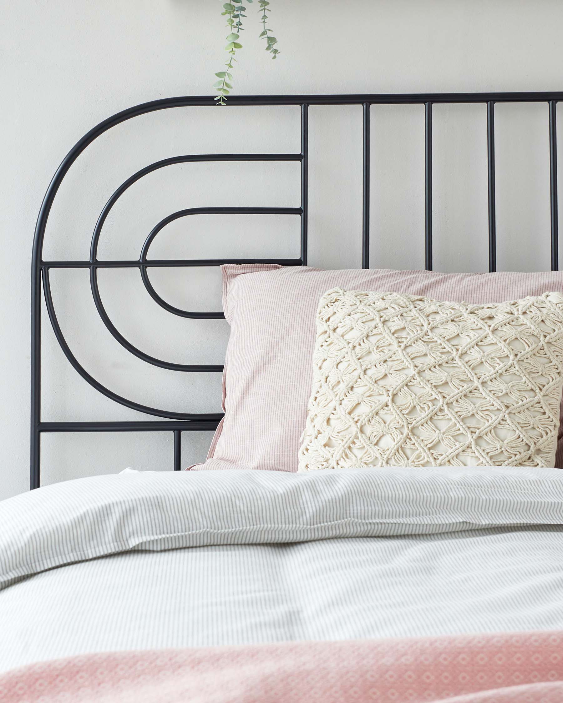 A modern metal bed frame with a geometric headboard pattern featuring circular and linear elements, in a black or dark finish. The bed is dressed with a textured white duvet cover, a pale pink sheet with a subtle pattern, and a decorative cream pillow with a knitted design. A hanging green plant adds a touch of nature to the scene.