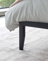 Close-up of a modern bed leg in black, showing a sleek and sturdy design supporting the corner of a bed with striped bedding. The bed stands on a plush, light-coloured carpet flooring, suggesting a sophisticated and contemporary bedroom setting.