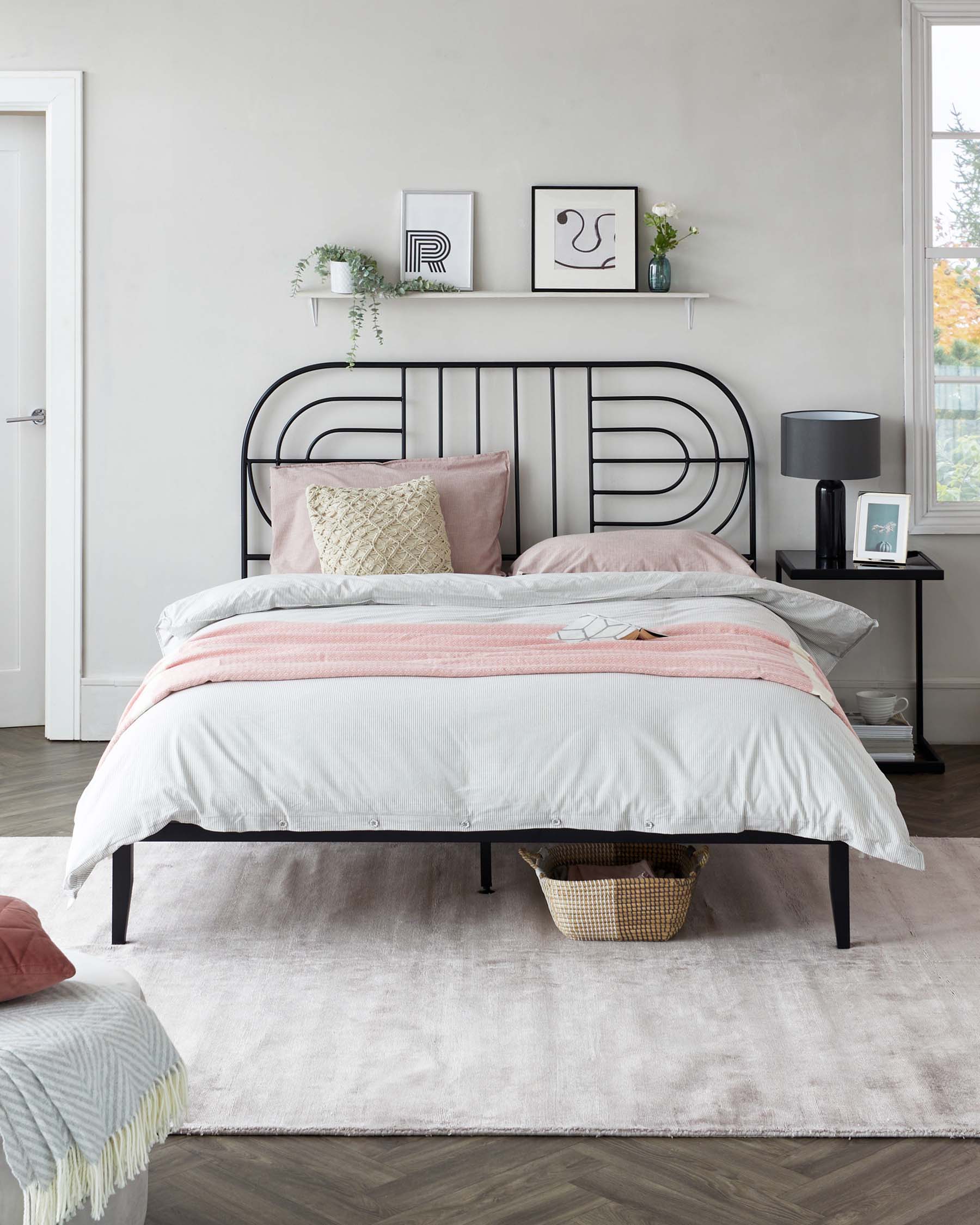 A contemporary-style bedroom featuring a metal frame bed with a minimalist headboard design, complemented by soft pink and grey bedding. Next to the bed stands a sleek black bedside table with a modern lamp, all anchored upon a plush, light grey area rug. A small wicker basket can be observed under the bed.