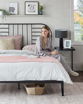 A contemporary bedroom featuring a stylish black metal bed frame with a decorative headboard, a small black bedside table with a sleek black lamp and picture frame, and a simple white floating shelf above the bed displaying artwork and a vase with flowers. A textured wicker basket is placed under the bed for additional storage. The bed is dressed in soft pastel linens, adding a cosy and modern feel to the room.