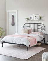 Modern minimalist bedroom with a black metal bed frame featuring a curved headboard, set against a light grey wall. The bed is dressed with white and blush pink bedding, accented by decorative pillows. A light pink area rug rests beneath the bed, and a white floating shelf above the headboard displays framed artwork. A woven basket sits at the bed's foot on the rug, softening the overall contemporary aesthetic.