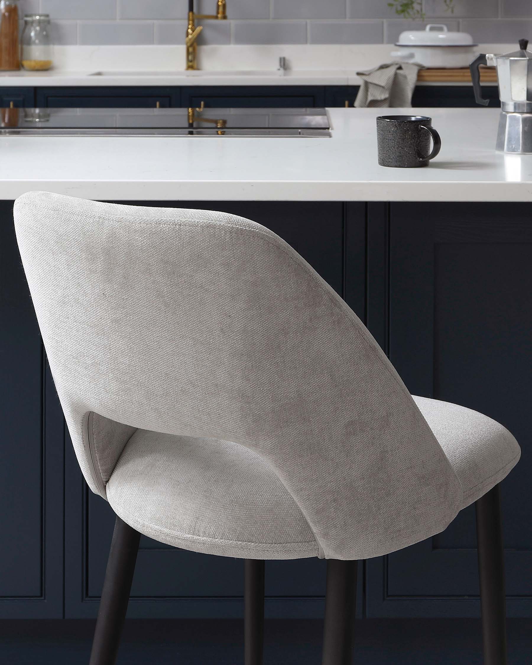 Modern light grey upholstered dining chair with a curved backrest and cushioned seat, mounted on sleek black metal legs. The chair is showcased in a stylish kitchen with dark blue cabinetry and white countertops.