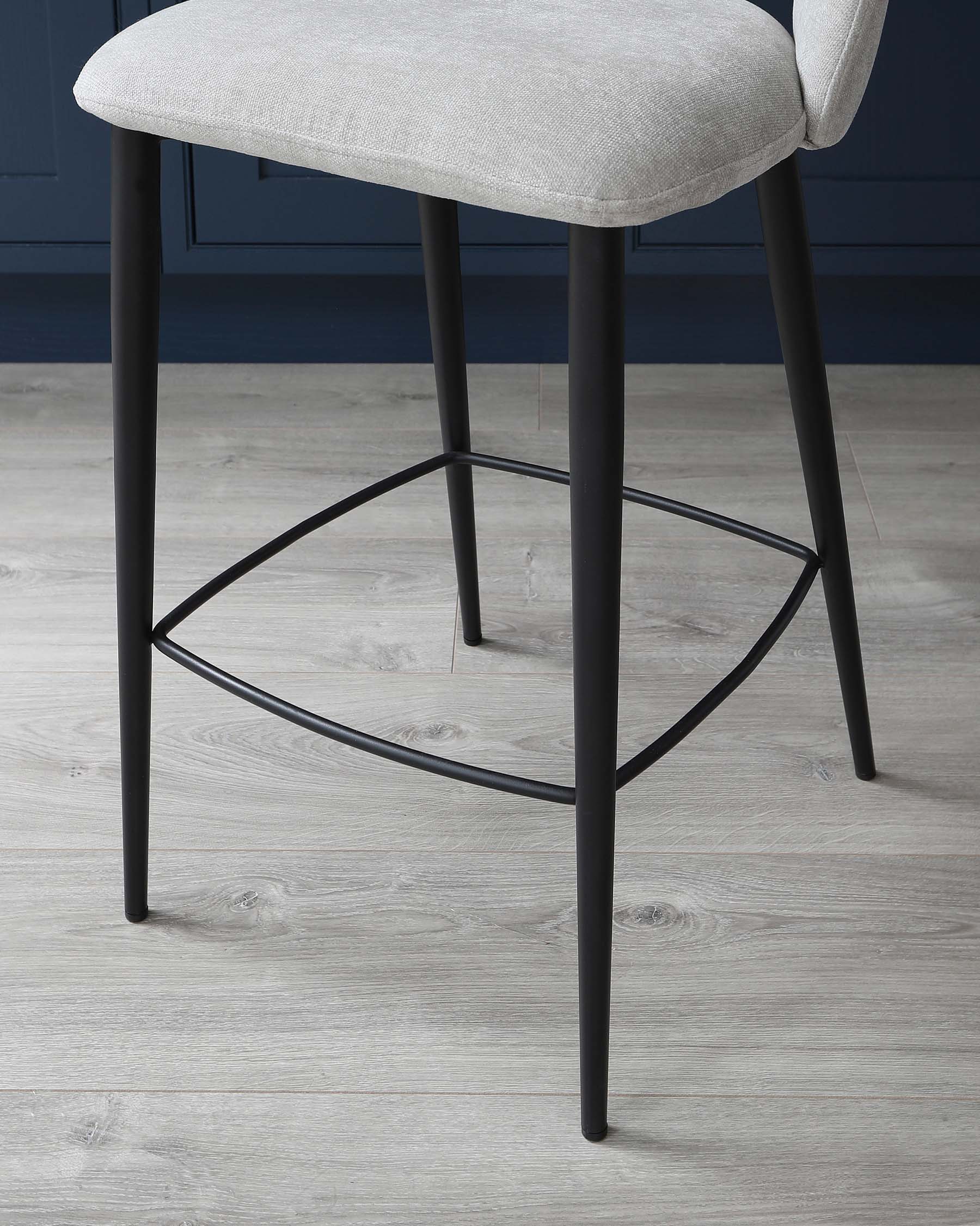 Modern minimalist style stool with a light grey fabric cushioned seat and sleek black metal legs, featuring a geometric footrest design.