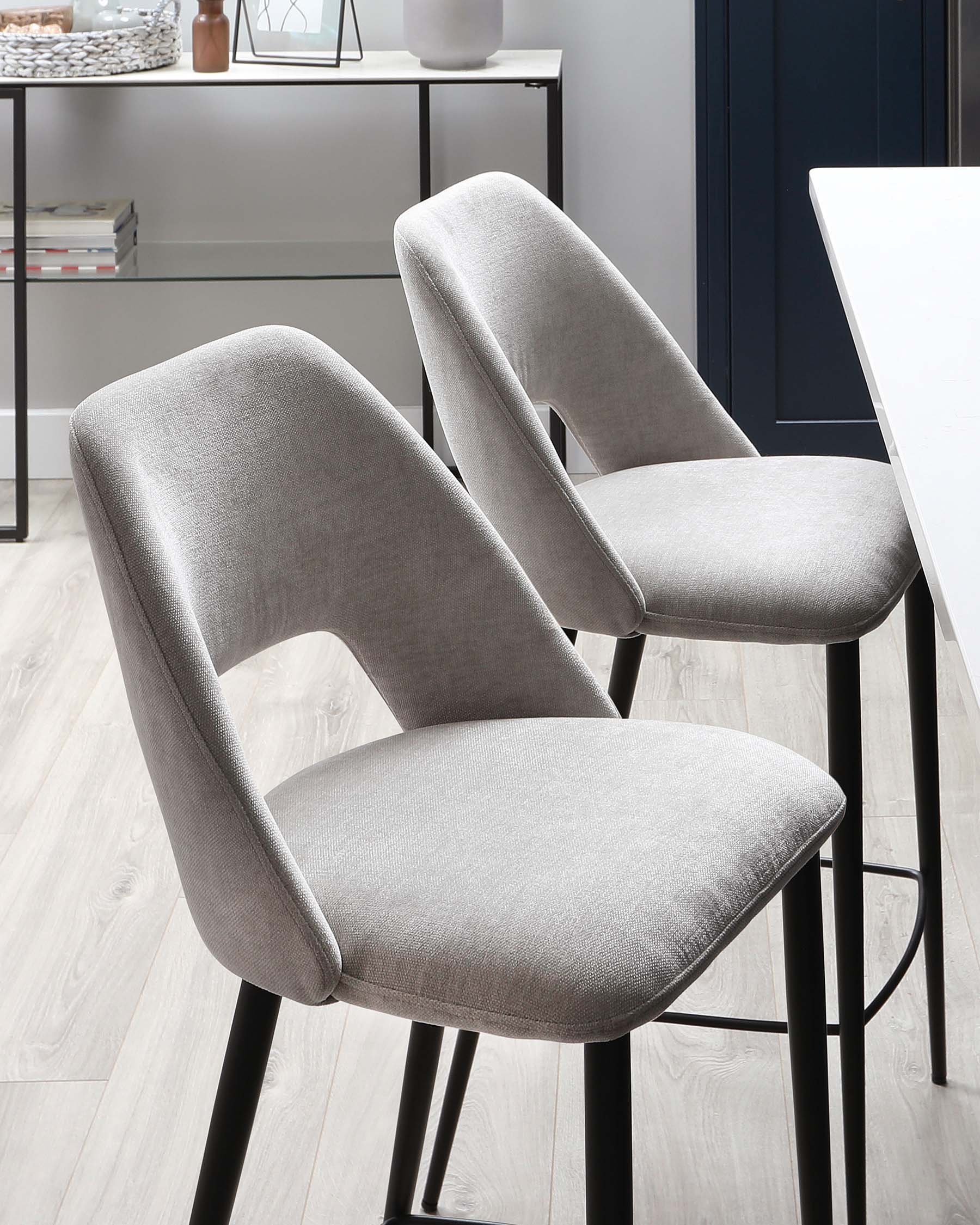 Two modern light grey upholstered dining chairs with curved backs and comfortable seat design, featuring sleek black metal legs. The chairs are positioned beside a white dining table, complementing a contemporary kitchen interior with a minimalist black shelving unit visible in the background.