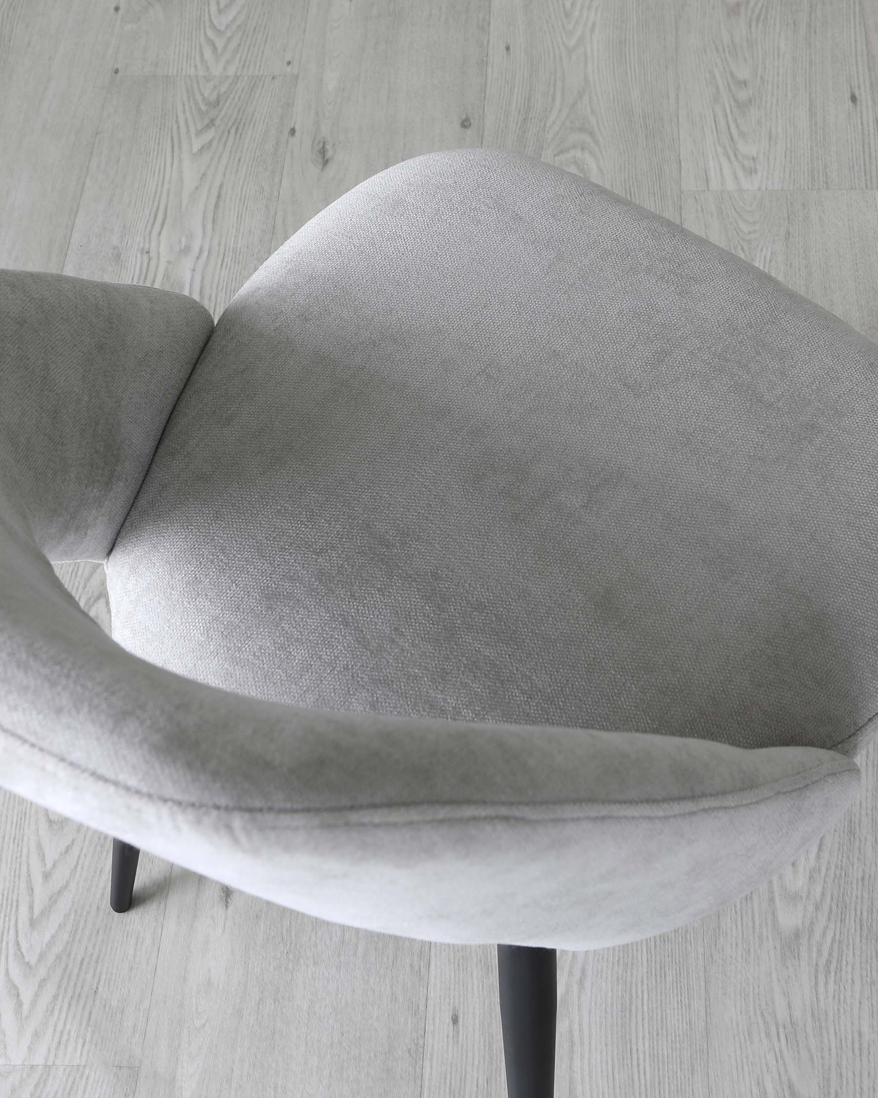 Modern light grey upholstered chair with a curved backrest and seat design, featuring simplistic black legs, set against a light wooden flooring background.