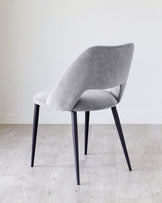 Modern light grey upholstered chair with a curved backrest and seat, featuring sleek black metal legs, positioned on a light wooden floor against a plain white background.