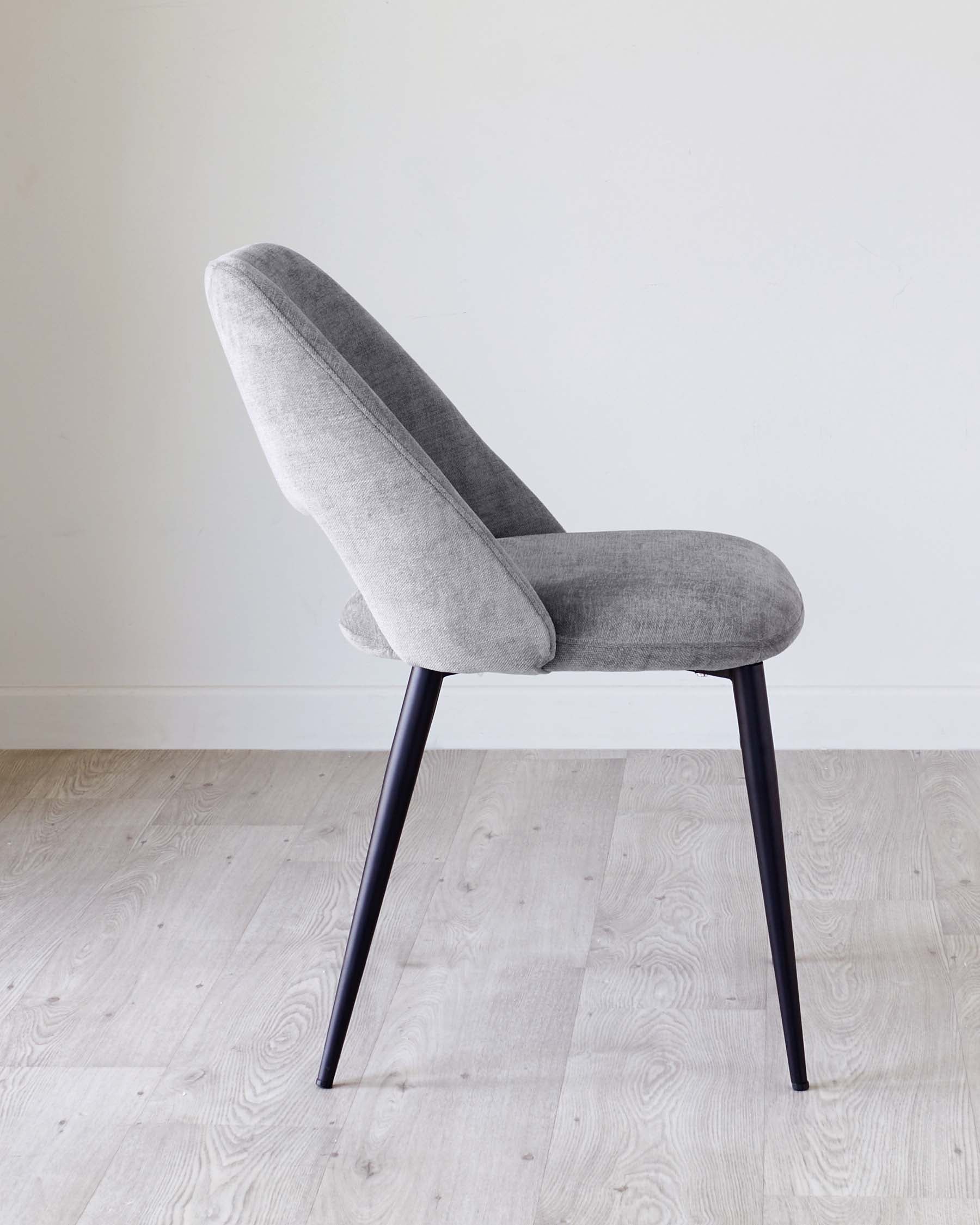 A modern, minimalist dining chair with a curved backrest and a comfortable, padded seat, both upholstered in light grey fabric. The chair features sleek, black metal legs that taper gently towards the floor. It is positioned on a pale wood laminate flooring against a plain white wall.