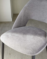 A modern chair featuring a curved backrest and a comfortably padded seat, upholstered in a light grey textured fabric, with slender dark metal legs, set against a light wooden floor and white walls background.