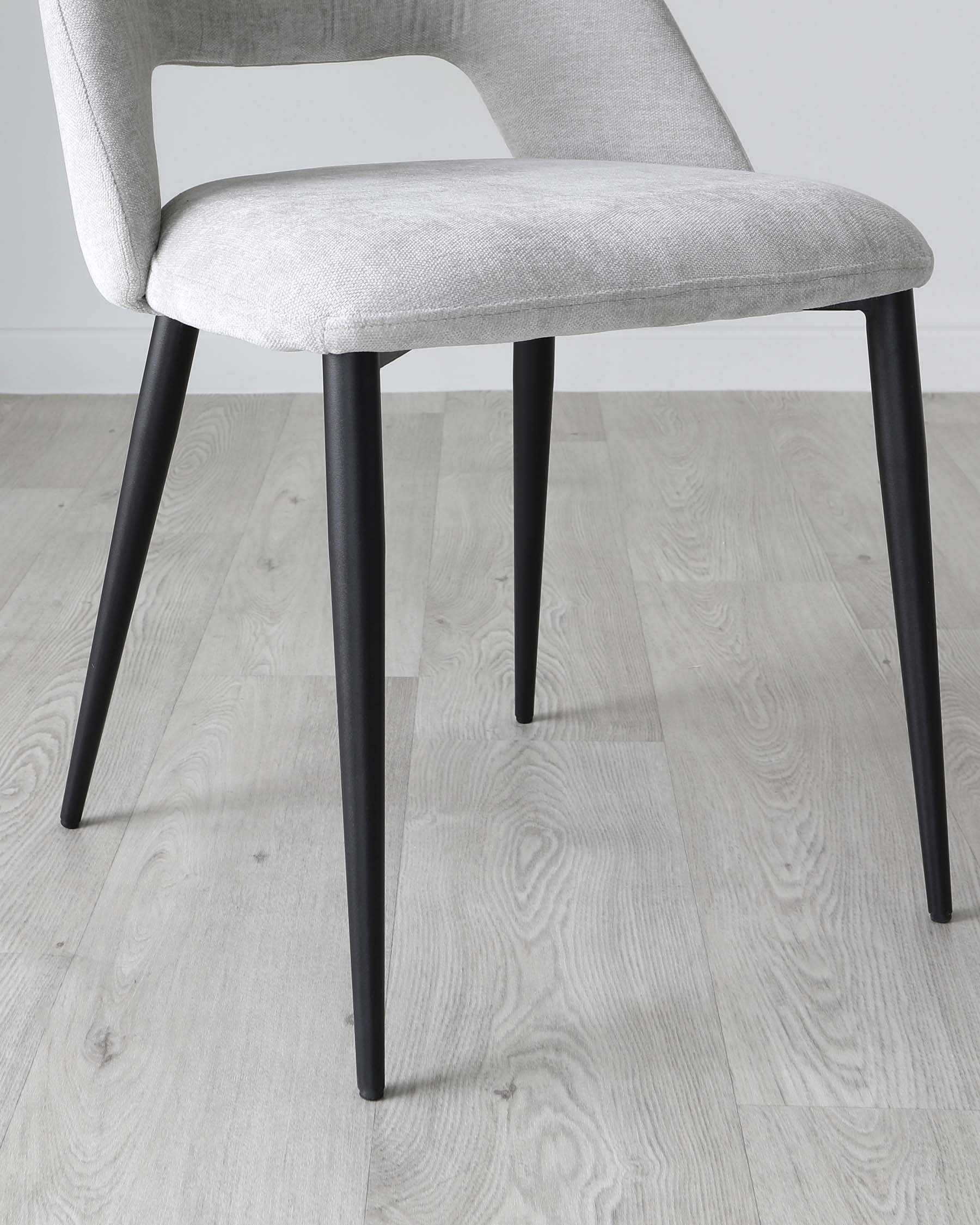 Modern minimalist dining chair with light grey fabric upholstery and sleek black metal legs, featuring a curved backrest for comfort, displayed on a light wood laminate floor.