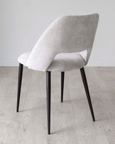 Modern light grey upholstered dining chair with a curved backrest and seat, featuring four slender black metal legs, positioned on a light wooden floor against a white wall.