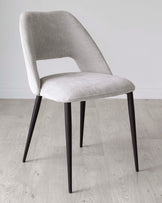 Modern upholstered dining chair with a curved backrest and seat in light grey fabric, featuring sleek black metal legs. The chair has a minimalist design with a cut-out detail on the lower back, positioned on a light wooden floor against a white background.