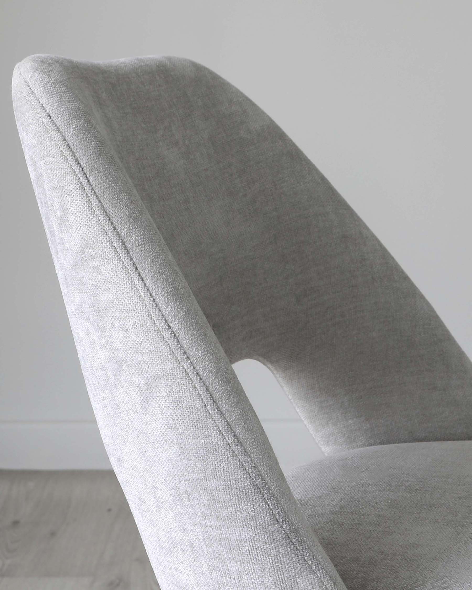 Close-up of a modern light grey fabric upholstered chair with a curved backrest and seat, showcasing the texture and contours of the design. The chair is against a neutral white wall on a light wooden floor, emphasizing its contemporary style and elegance.