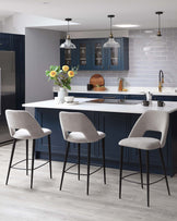 Three modern style bar stools with light grey upholstery and black metal legs are positioned around a white kitchen island. The stools feature a sleek design with a slight backrest and integrated footrests for comfort.