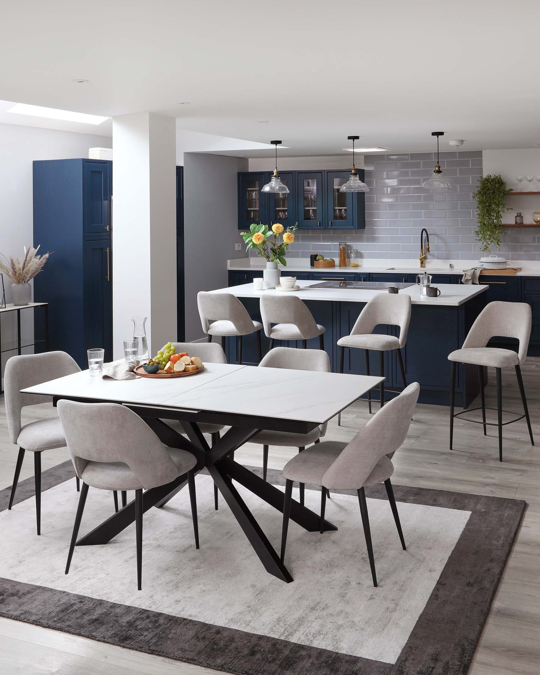 Modern dining room with a white rectangular table featuring a marble-look top and black angular legs. Six contemporary grey upholstered chairs with black legs surround the table. A grey and white area rug underlays the dining set. The scheme coordinates with the kitchen bar stools and cabinetry in the background.