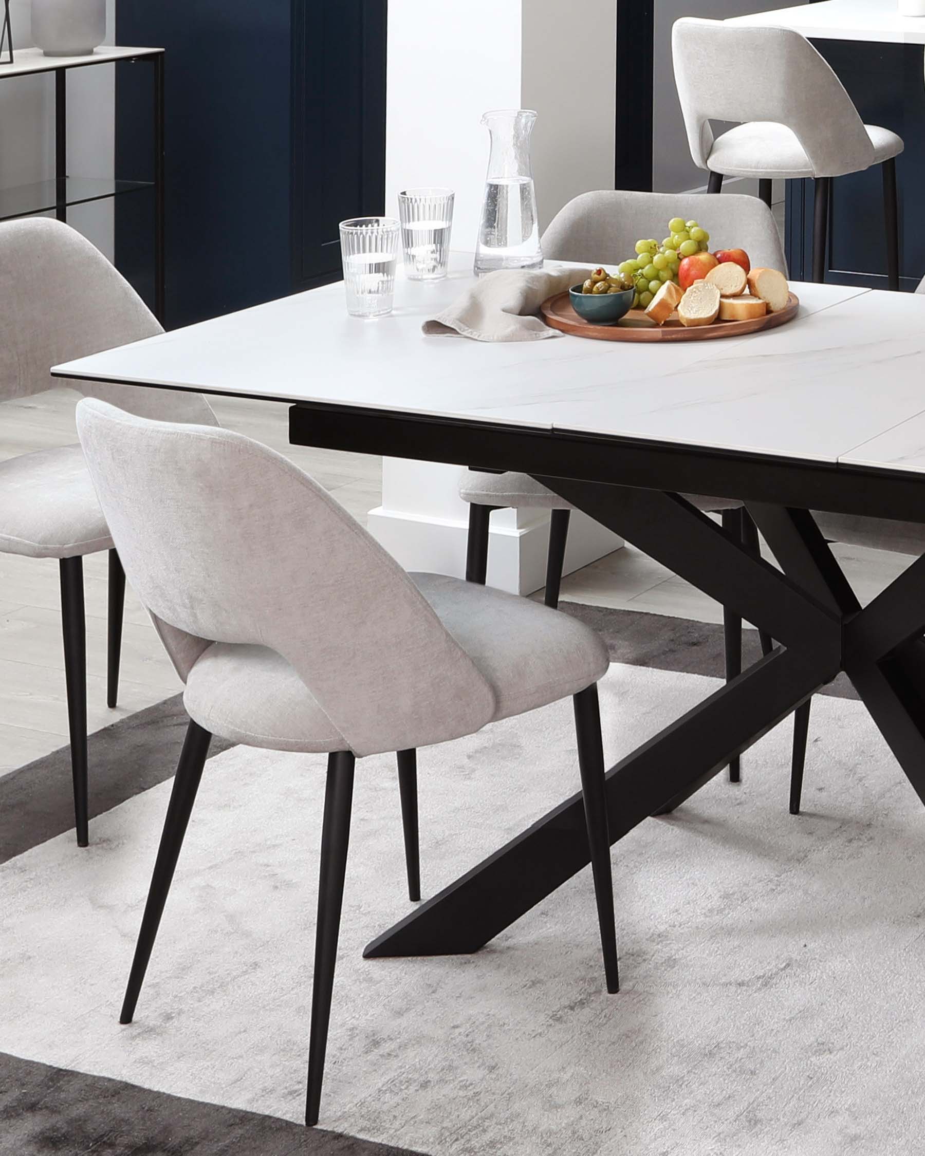 Modern dining room furniture featuring a rectangular white marble top table with black angular legs, paired with light grey upholstered chairs with sleek black frames. A matching sideboard is partially visible in the background.