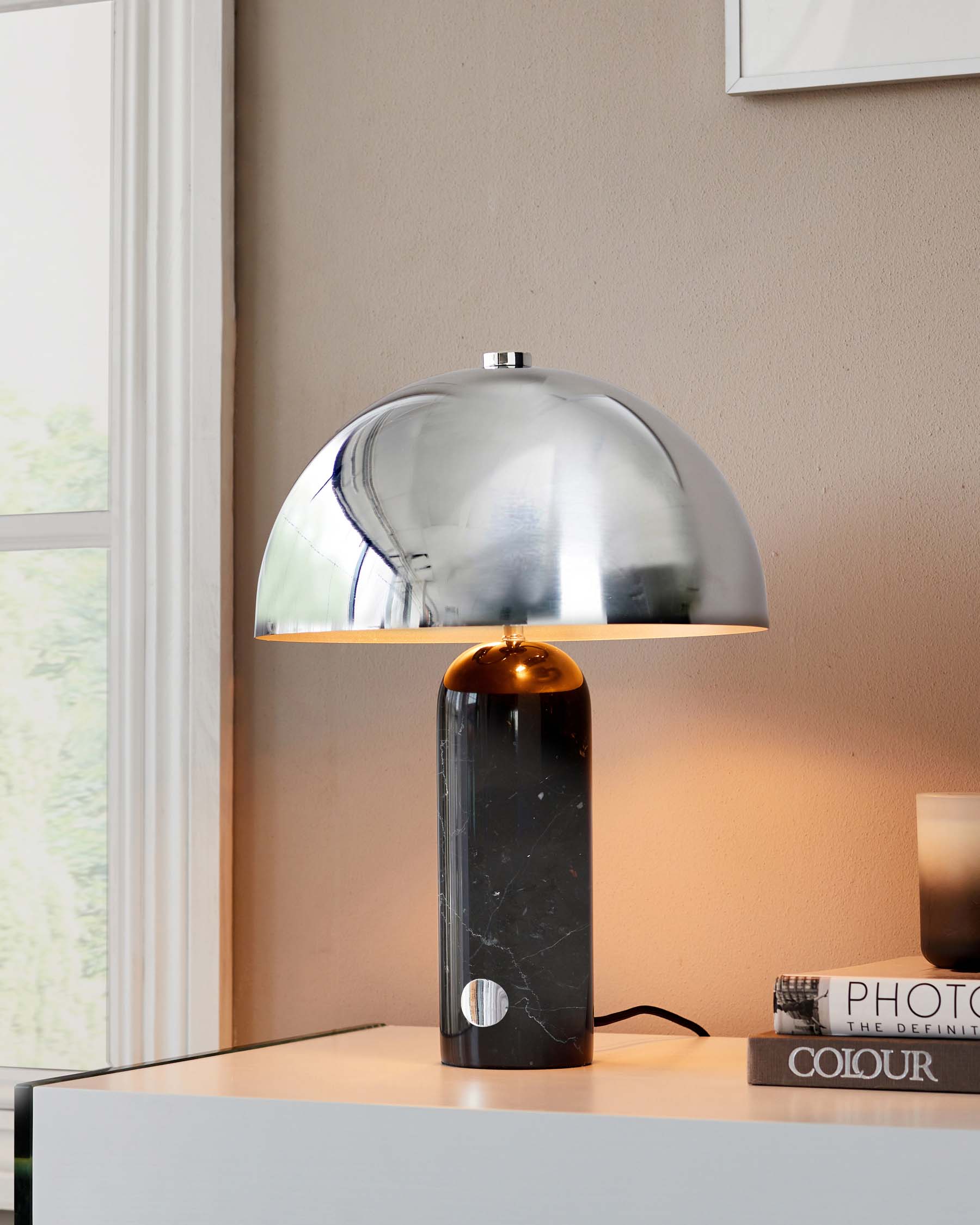 A contemporary table lamp with a glossy chrome dome shade and a cylindrical marble base, positioned on a sleek white surface, with books and a window providing natural light in the background.