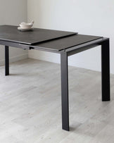 Modern minimalist black extendable dining table with a matte finish and square legs, partially extended with a set of white dishes on top, placed on a light hardwood floor against a white wall.