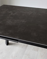 Modern minimalist coffee table with a dark stone-like finish and sleek black metal legs, positioned on a pale wooden floor against a plain white background.