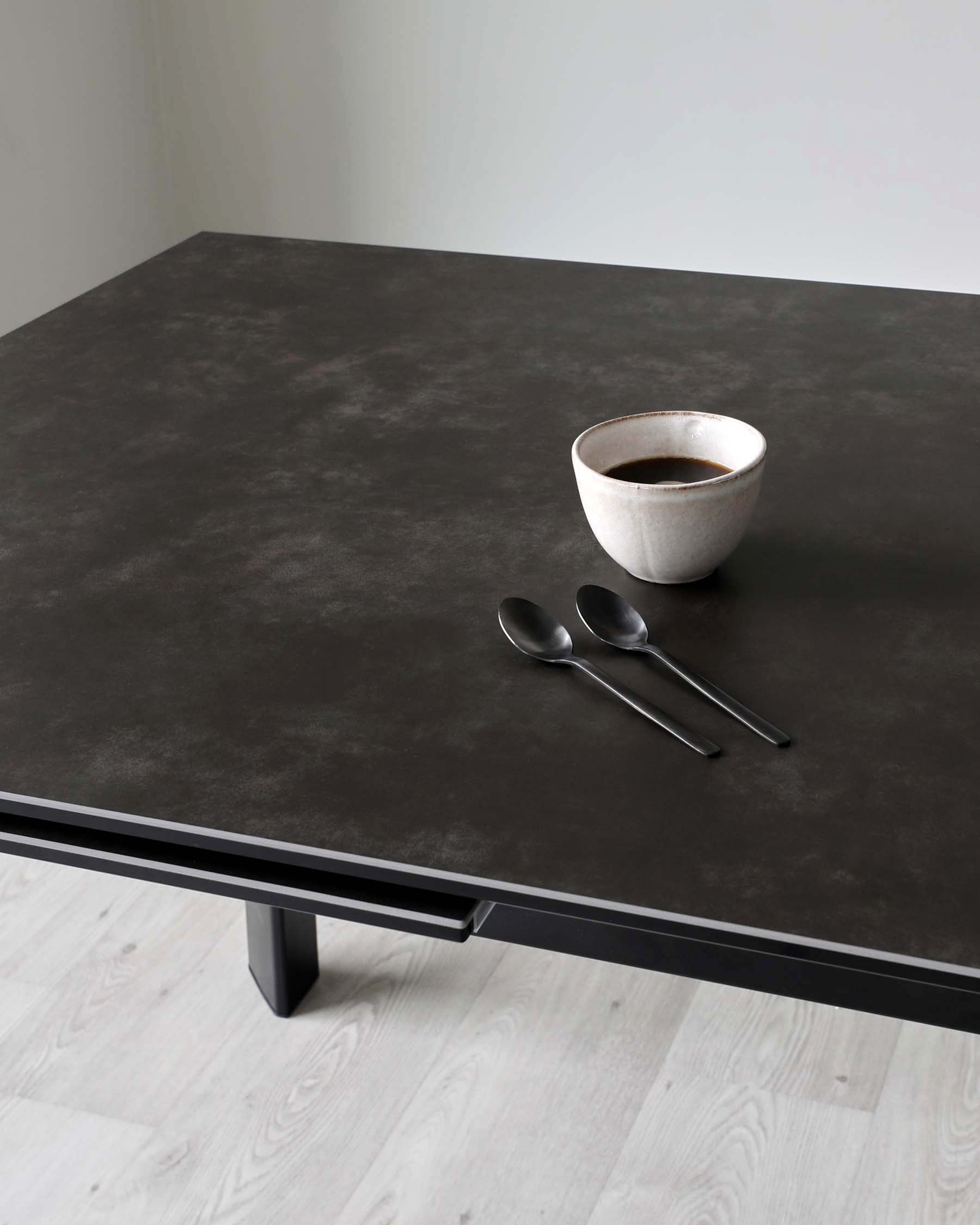 Modern minimalist dark brown dining table with a matte finish and sturdy rectangular legs, complemented by a simple ceramic bowl and a pair of spoons on top.