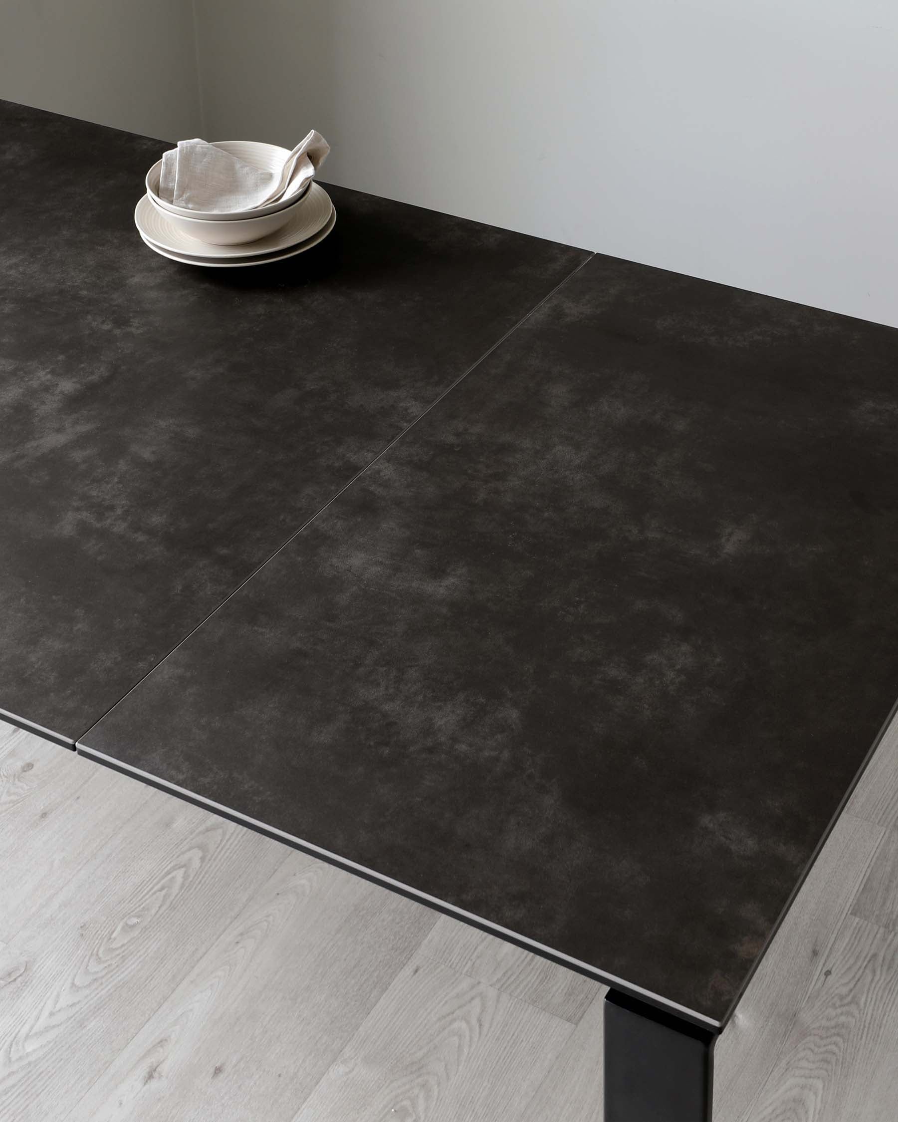 Minimalist matte black table with a stack of off-white plates on top, set against a light wood floor and pale grey wall.