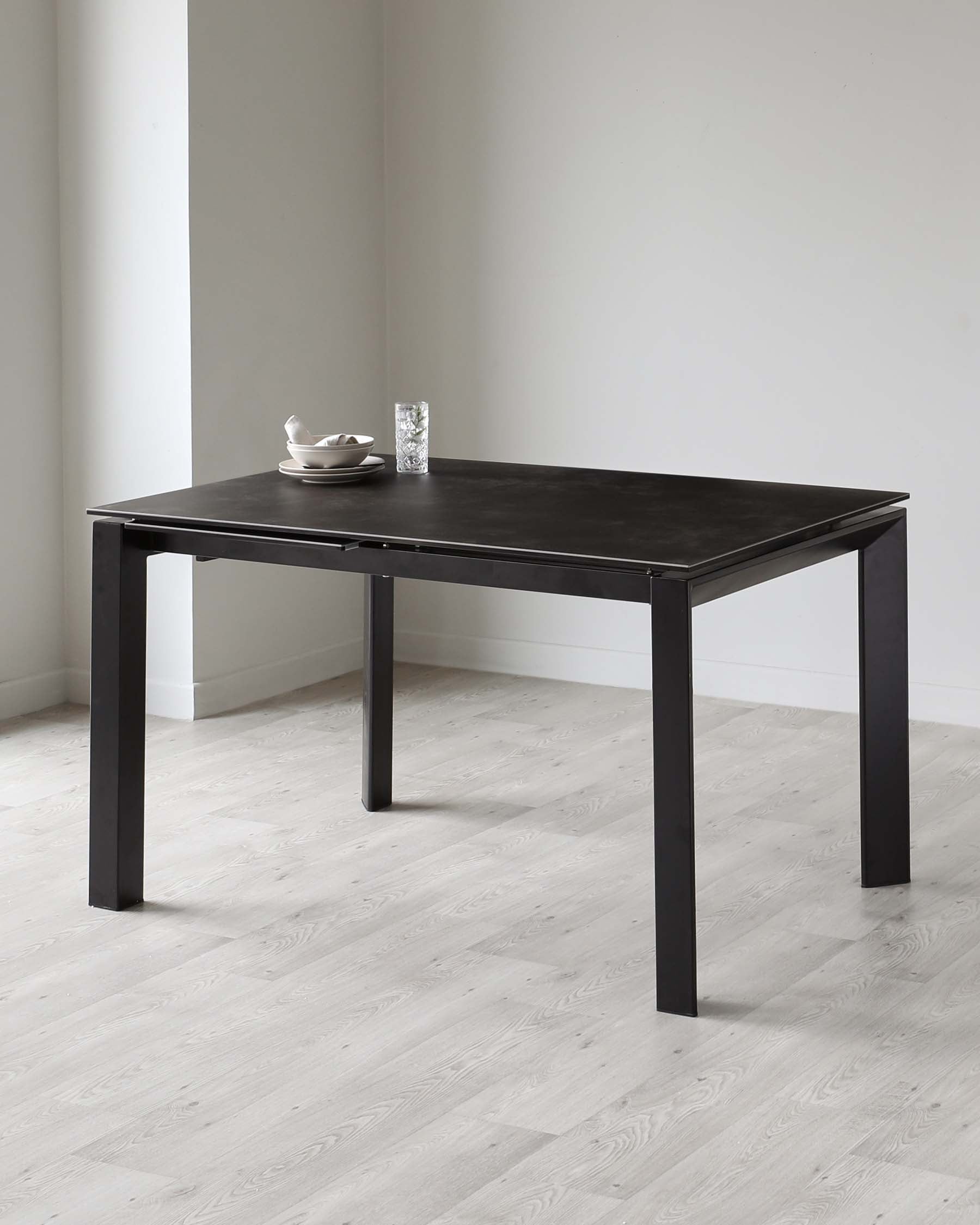 A modern, minimalist black dining table with a square top and four straight legs. The tabletop is accessorized with a white bowl and a glass tumbler, indicating its use in a dining setting. The table is placed on a light hardwood floor against a neutral wall, emphasizing its sleek design.