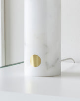 Elegant white marble cylindrical lamp base with a brass accent detail, paired with a transparent glass surface that appears to be the edge of a larger glass table.