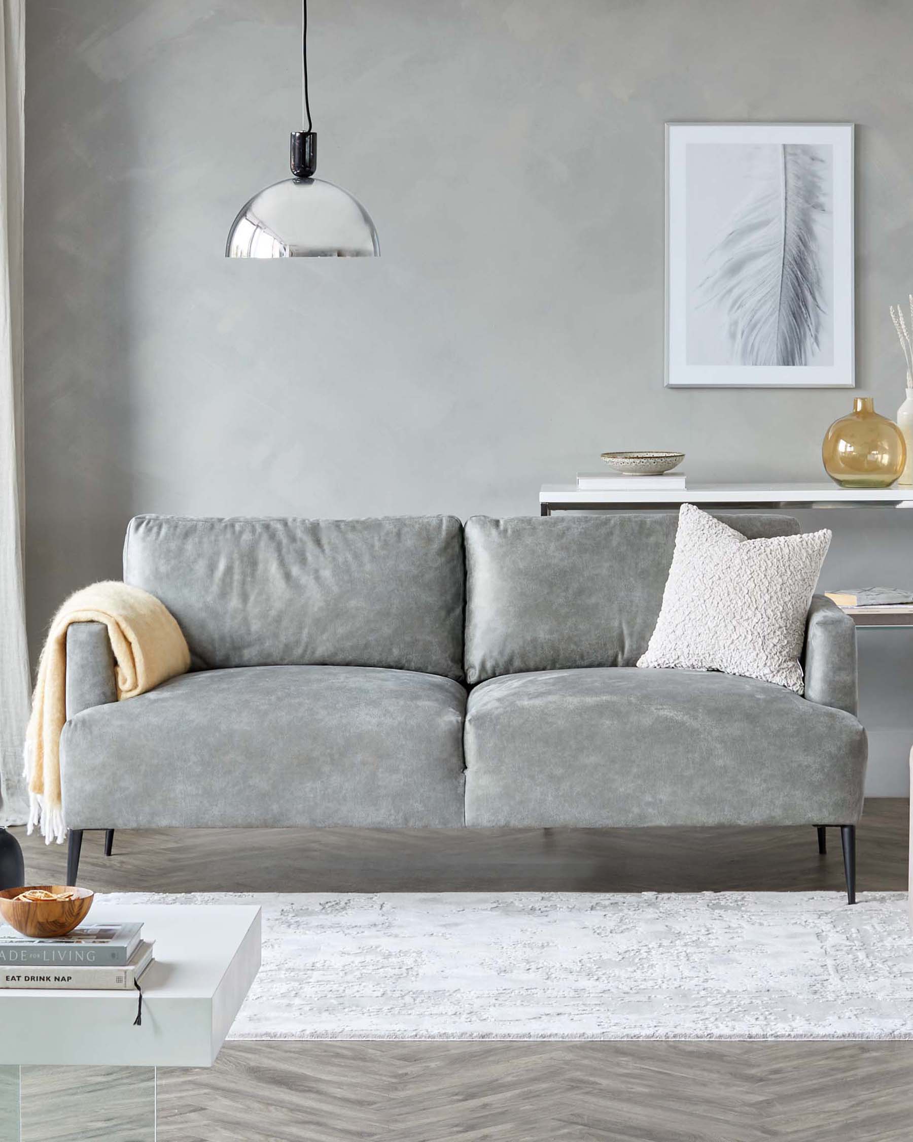 Modern living room featuring a plush two-seater grey velvet sofa with two cushions, sleek metal legs, complemented by a white coffee table with books and a wooden bowl atop a textured white area rug. A minimalist white console table with decorative vases and bowl against a grey wall, and a contemporary pendant light completes the setting.