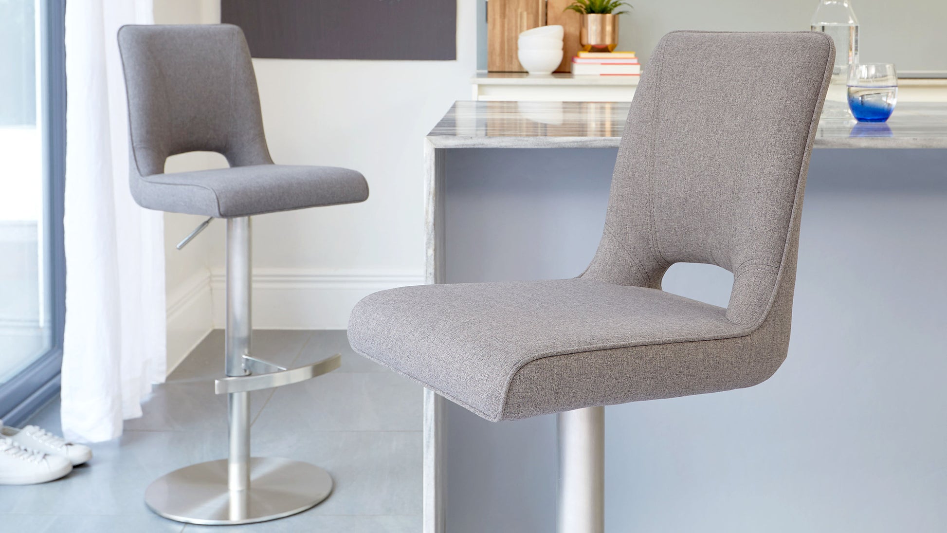 Two contemporary grey fabric bar stools with backrests, featuring clean lines, an L-shaped cut-out at the seat base, and brushed metal pedestal bases with footrests and adjustable height settings.