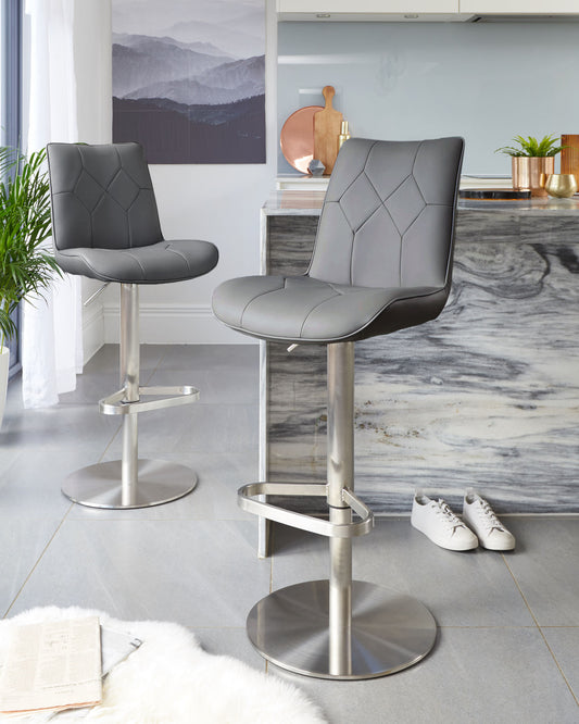 A pair of modern bar stools with chrome pedestals, featuring adjustable height, a footrest, and quilted faux leather seats in a neutral grey tone, suitable for contemporary kitchen or bar areas.