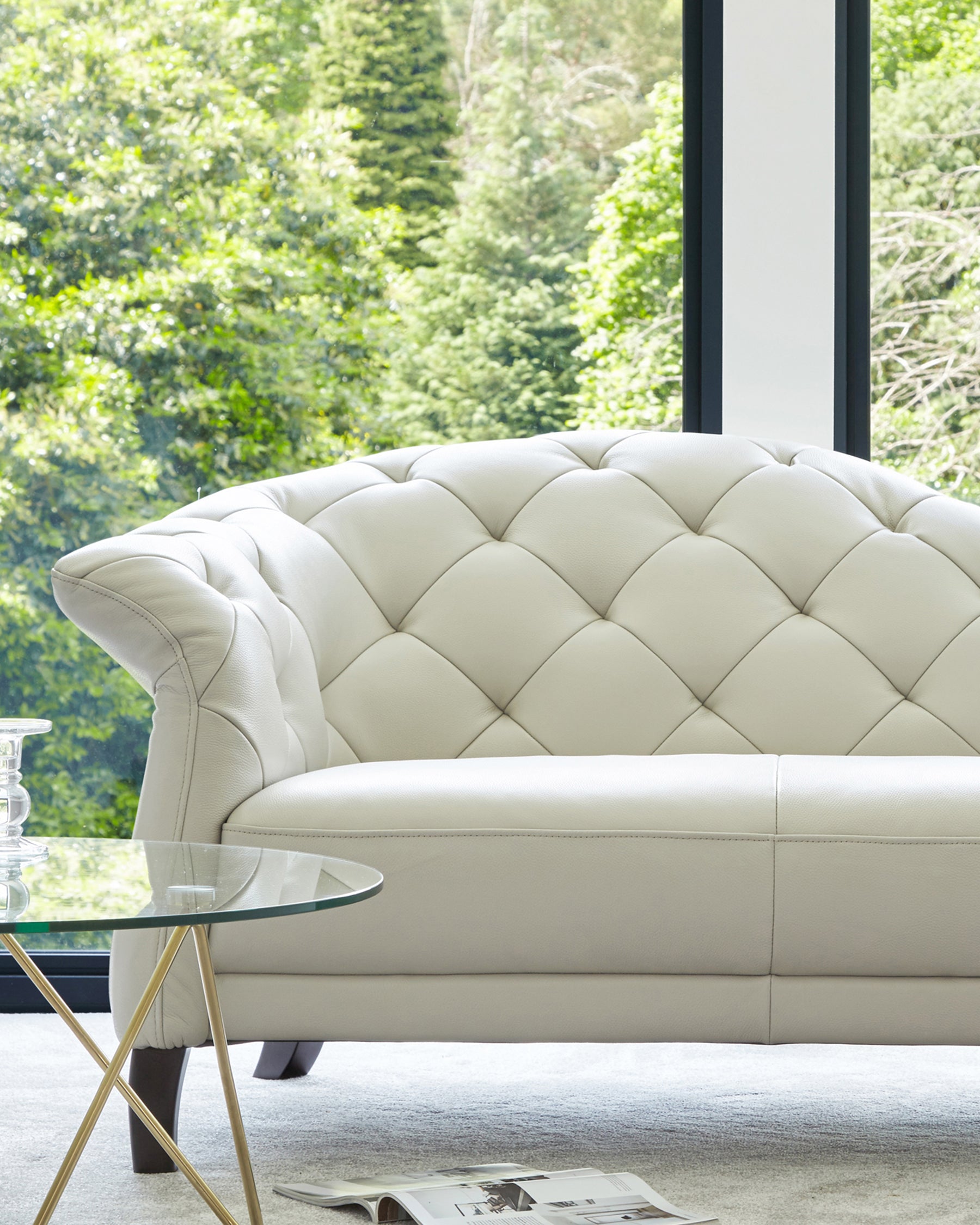 Elegant white tufted sofa paired with a modern glass top coffee table featuring a sleek gold base, set against a lush backdrop.