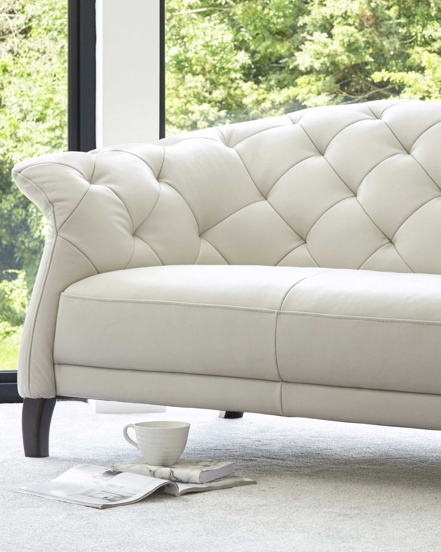 A sleek, cream-colored tufted sofa with elegant curves, resting on dark wooden legs, accompanied by a decorative cup and magazines.