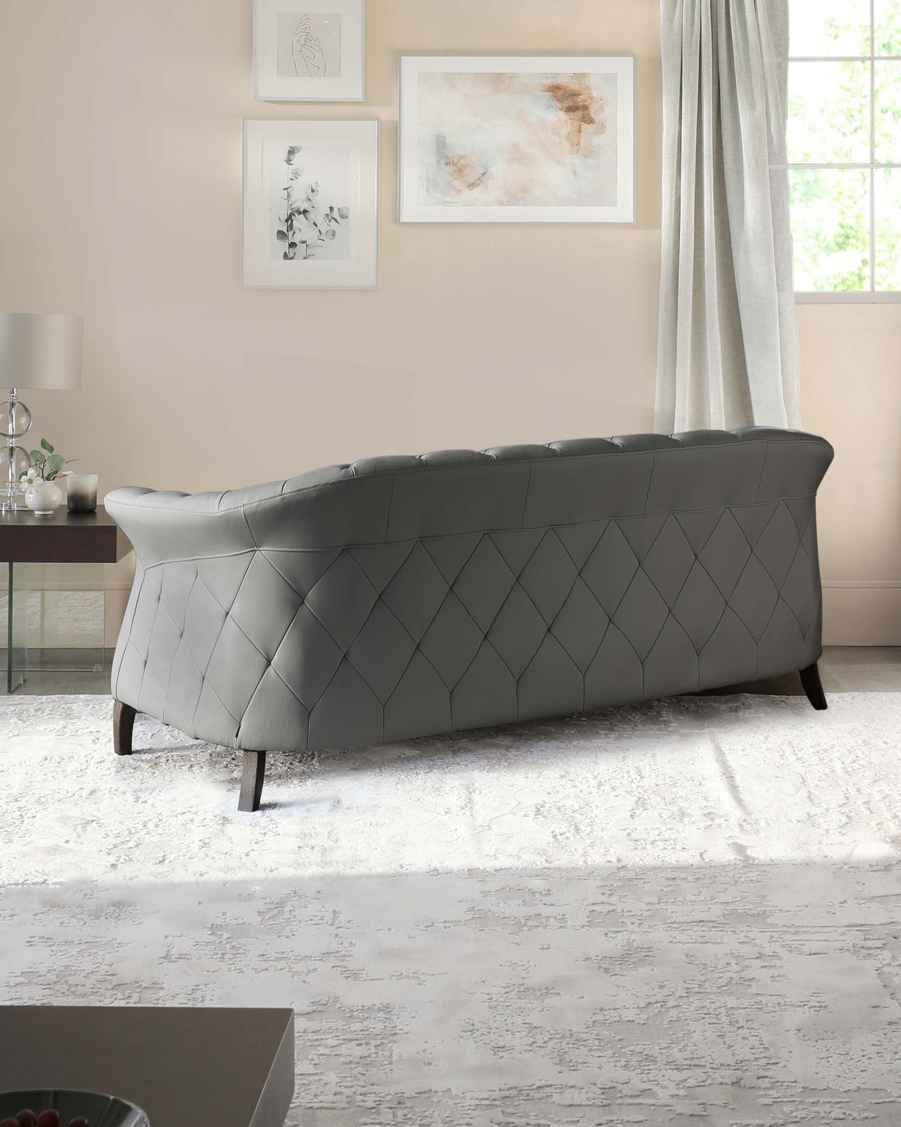 Elegant tufted grey sofa with a curved backrest featuring geometric stitching patterns, placed on a textured white area rug in a tastefully decorated room with a light neutral colour palette.